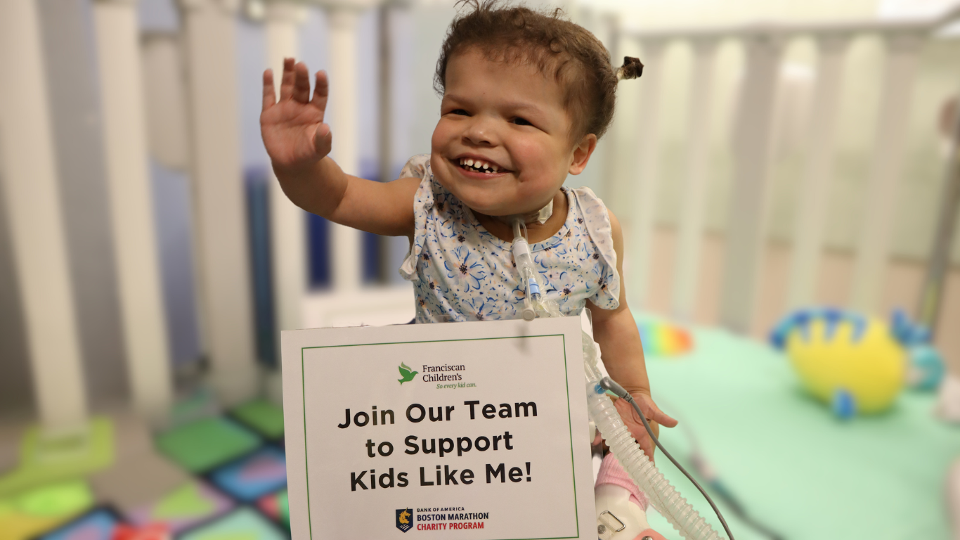 Young child with a tracheostomy tube smiles and waves. In front of her is a sign that reads, "Join Our Team to Support Kids Like Me!"